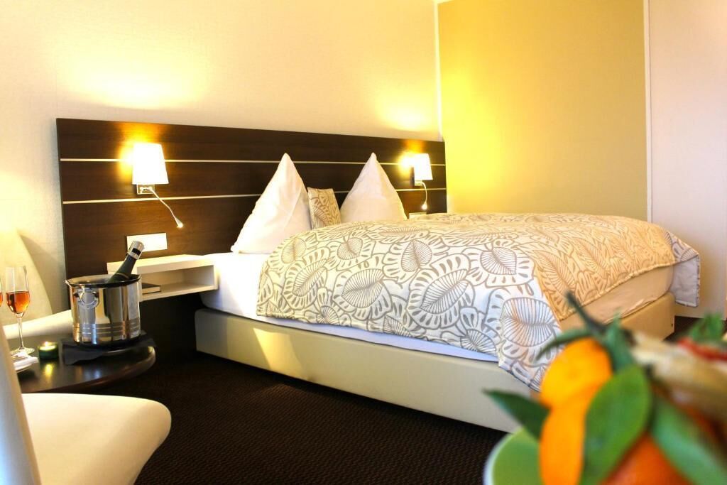 A hotel room with a bed, bedside lamps, a nightstand with a bottle and glasses, and a small table with a candle.