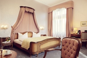 A four-poster canopy bed with a wooden frame and white bedding, a round wooden table with a cup and saucer, and a tufted armchair. - Schlosshotel Rettershof (Kelkheim)