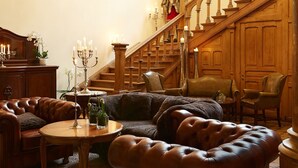 A spacious living room with a large brown leather sofa, a wooden coffee table, and a chandelier. A staircase with wooden railings leads to an upper floor. - Romantik Hotel Schloss Rettershof (Kelkheim)