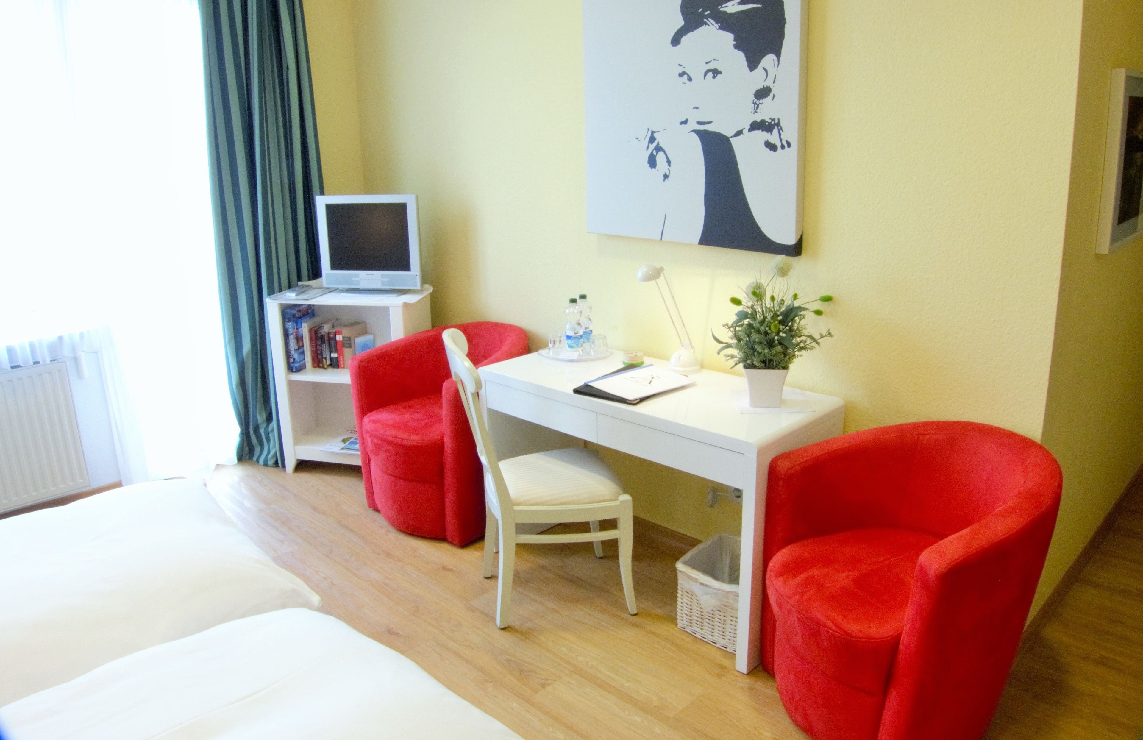 Ein Hotelzimmer mit einem Bett, einem Schreibtisch mit Computer, zwei roten Stühlen, einem weißen Stuhl, einer kleinen Pflanze und einem Bild an der Wand.