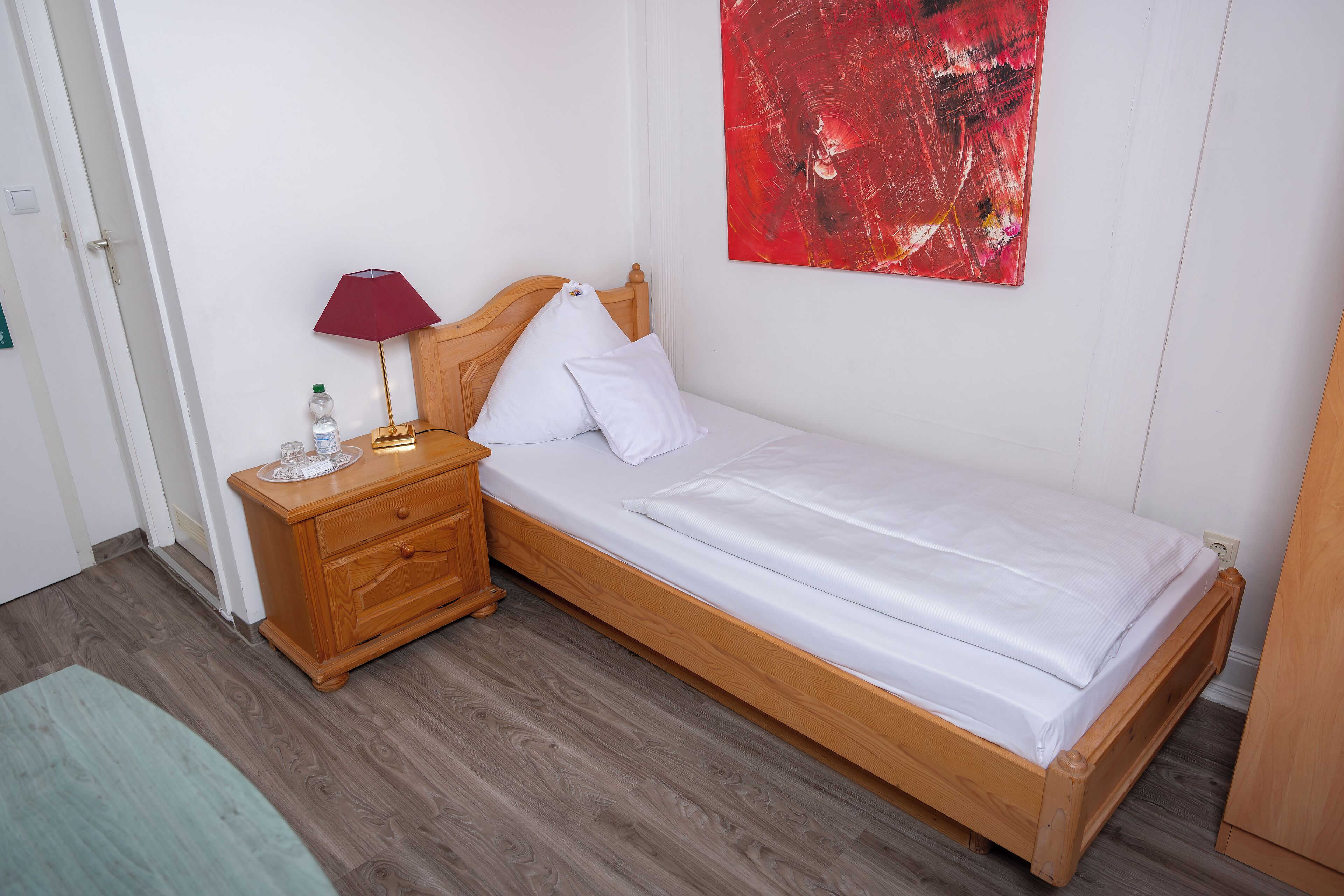 A single bed with a wooden frame, a wooden nightstand with a lamp, and a red abstract painting on the wall.