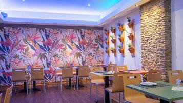 Un restaurante con mesas y sillas de madera, un mural tropical colorido y plantas en macetas sobre una pared de piedra.