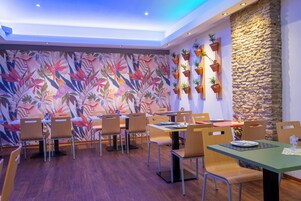 Un restaurante con mesas y sillas de madera, un mural tropical colorido y plantas en macetas sobre una pared de piedra.