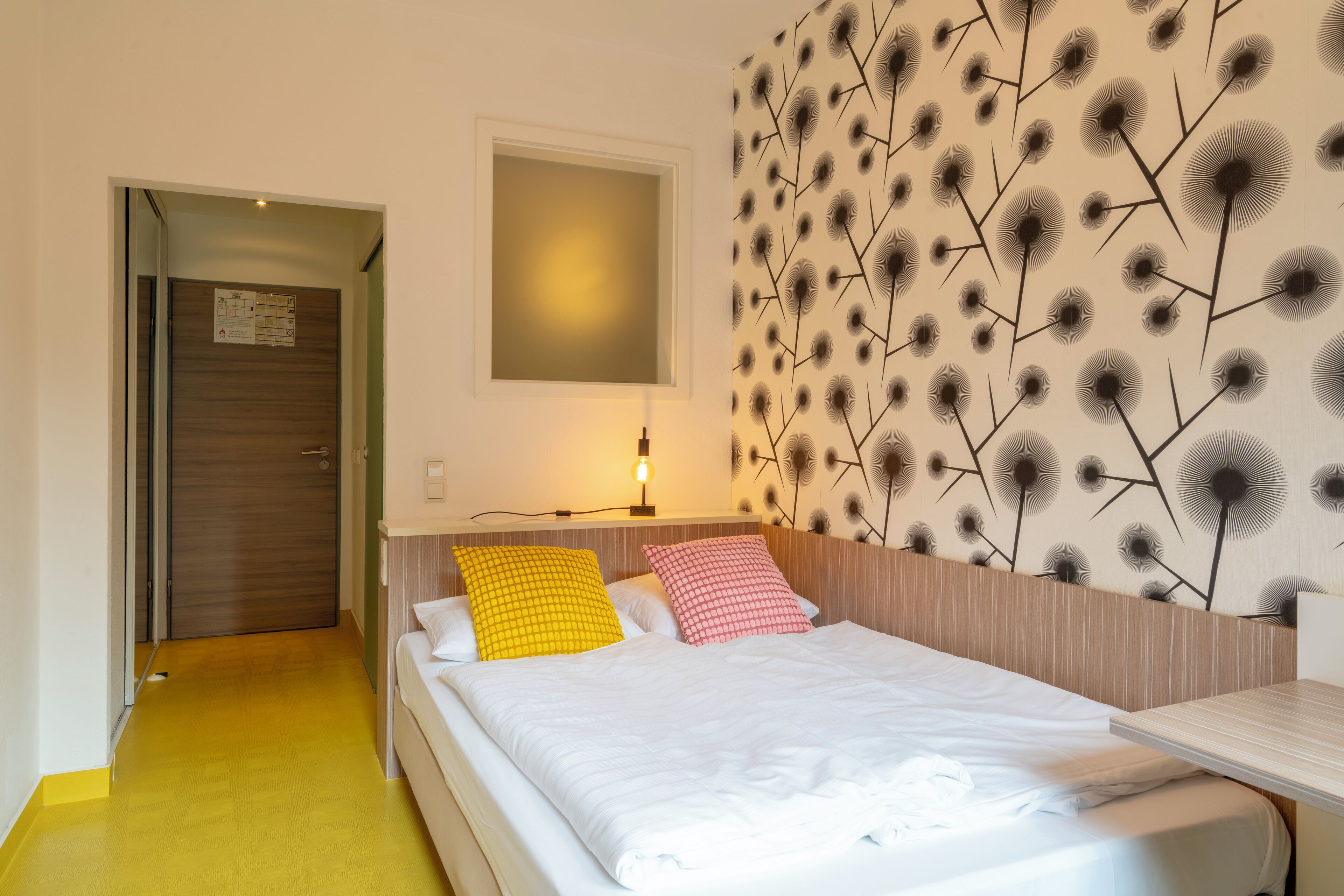 A hotel room with a bed, a bedside table, a wall with a leaf pattern, and a door.