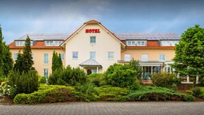 Ein Hotel mit zentralem Eingang, zahlreichen Fenstern und einem gepflegten Garten mit verschiedenen Sträuchern und Bäumen.