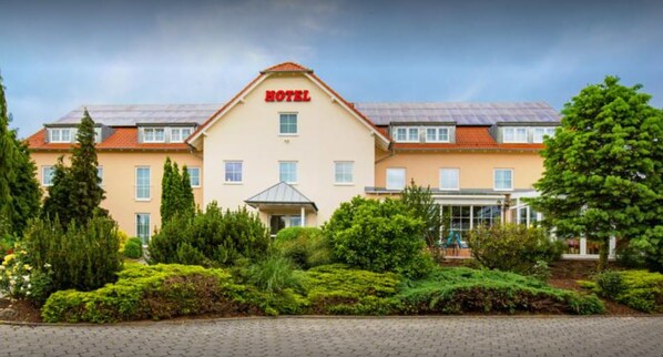 Un hotel con entrada central, múltiples ventanas y un jardín bien cuidado con arbustos y árboles variados.