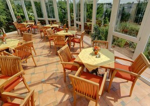 Un patio al aire libre con mesas y sillas de madera, rodeado de grandes ventanales con vistas a zonas verdes.