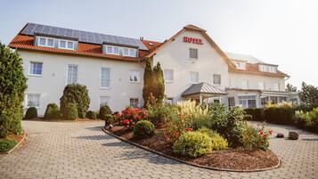 A hotel with a solar panel roof, a garden with red flowers, and a paved driveway.