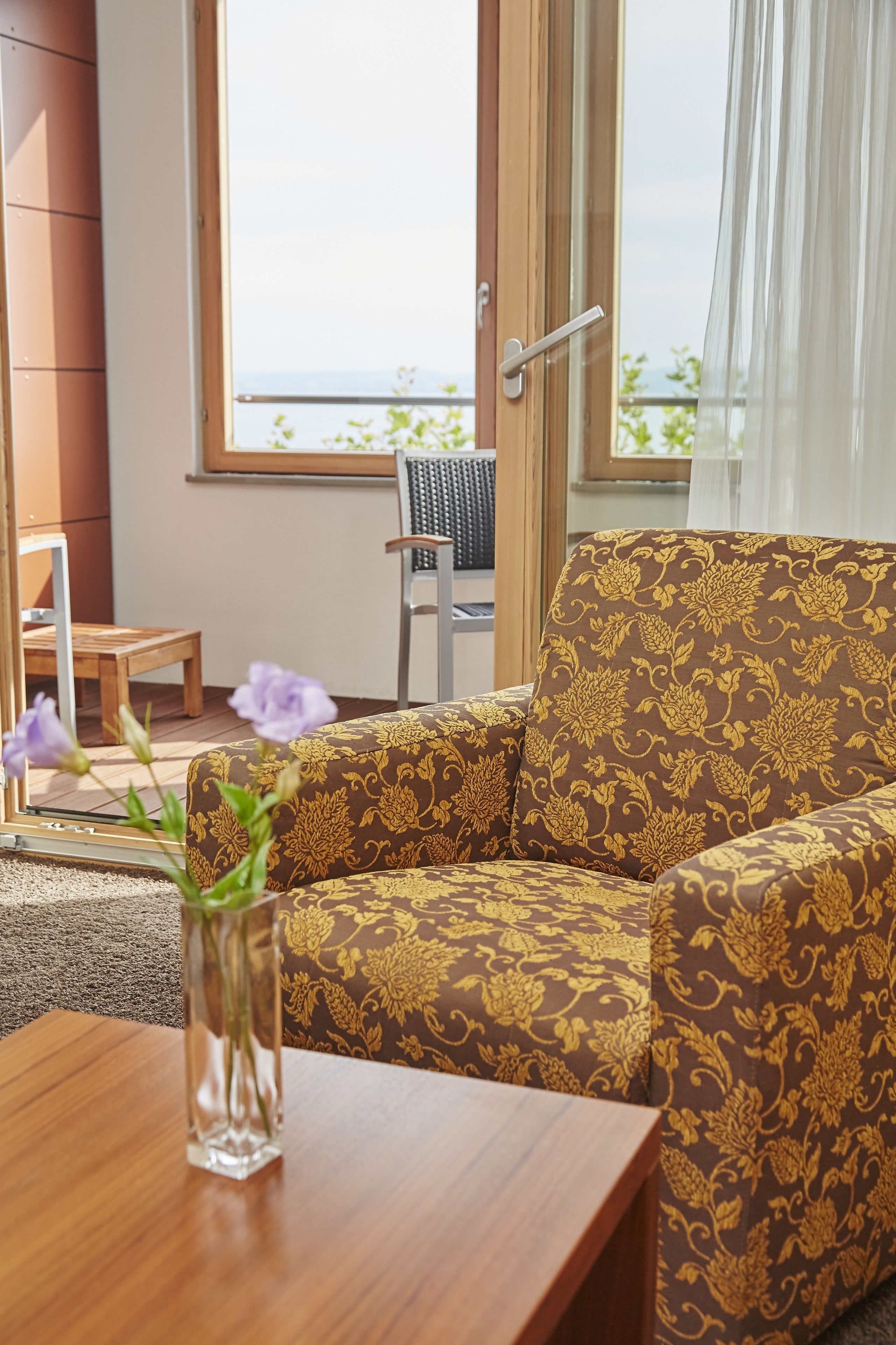 Ein Wohnzimmer mit geprägter Couch, einem Holztisch mit Blumenvase, einem Sessel und Blick ins Freie durch ein Fenster.