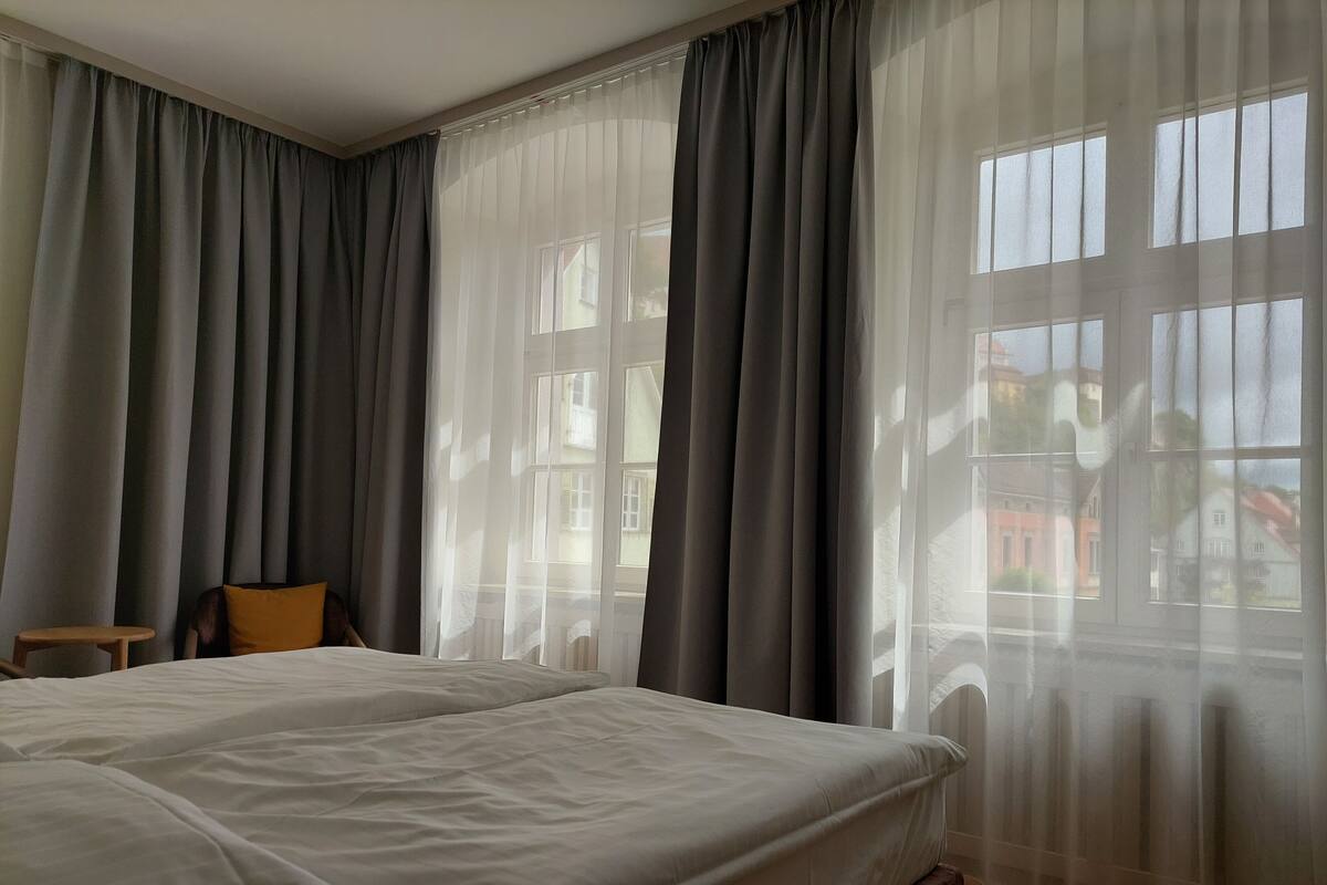 Hotelkamer met een groot bed, twee ramen met dunne gordijnen, een klein tafeltje en uitzicht op gebouwen.
