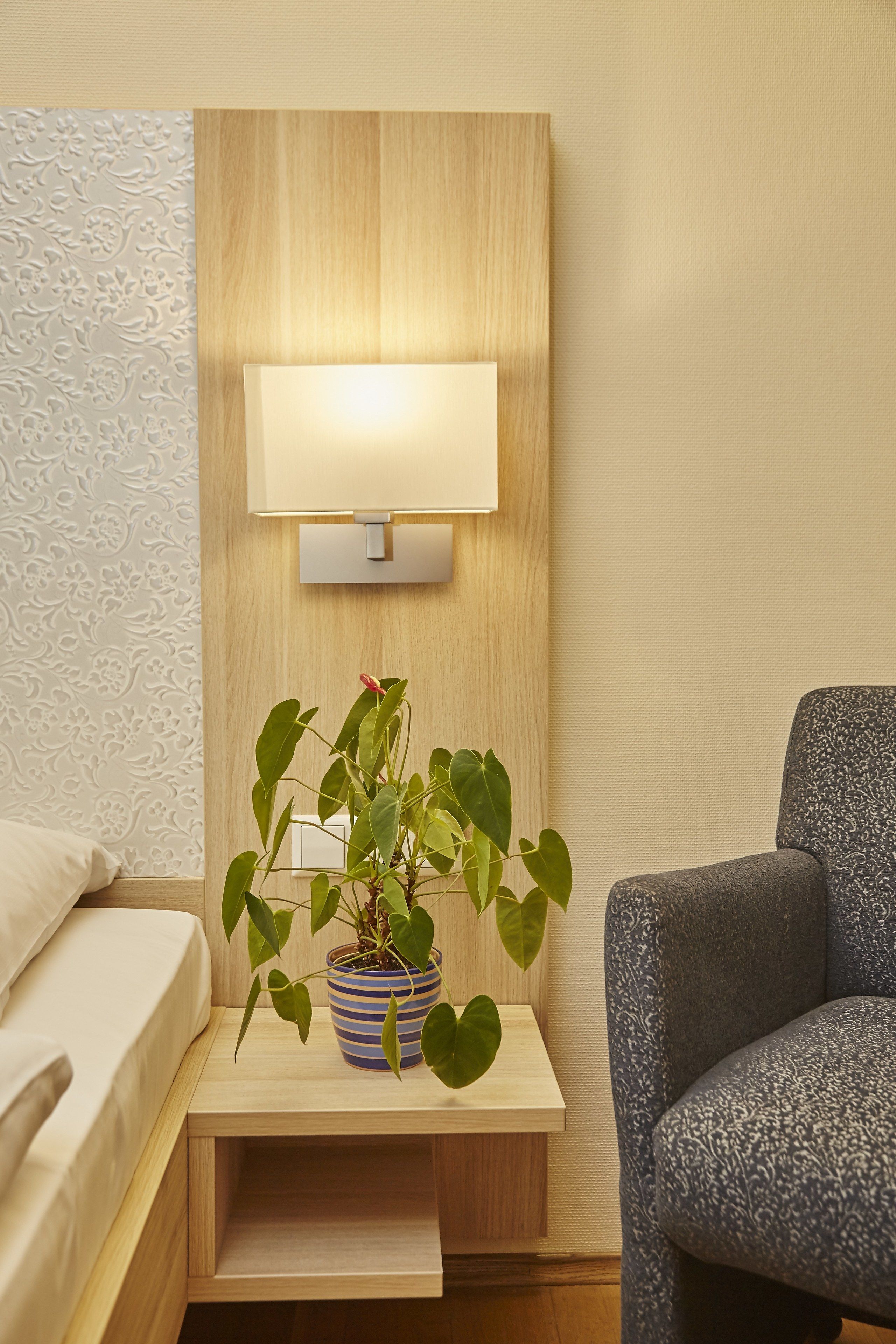 Habitación de hotel con una cama, una mesita de noche con lámpara, una planta en maceta y un sillón con estampado.