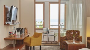 Une chambre d’hôtel avec un lit, un bureau équipé d’un ordinateur, un fauteuil jaune, un fauteuil à motifs et une vue sur la mer par de grandes fenêtres.