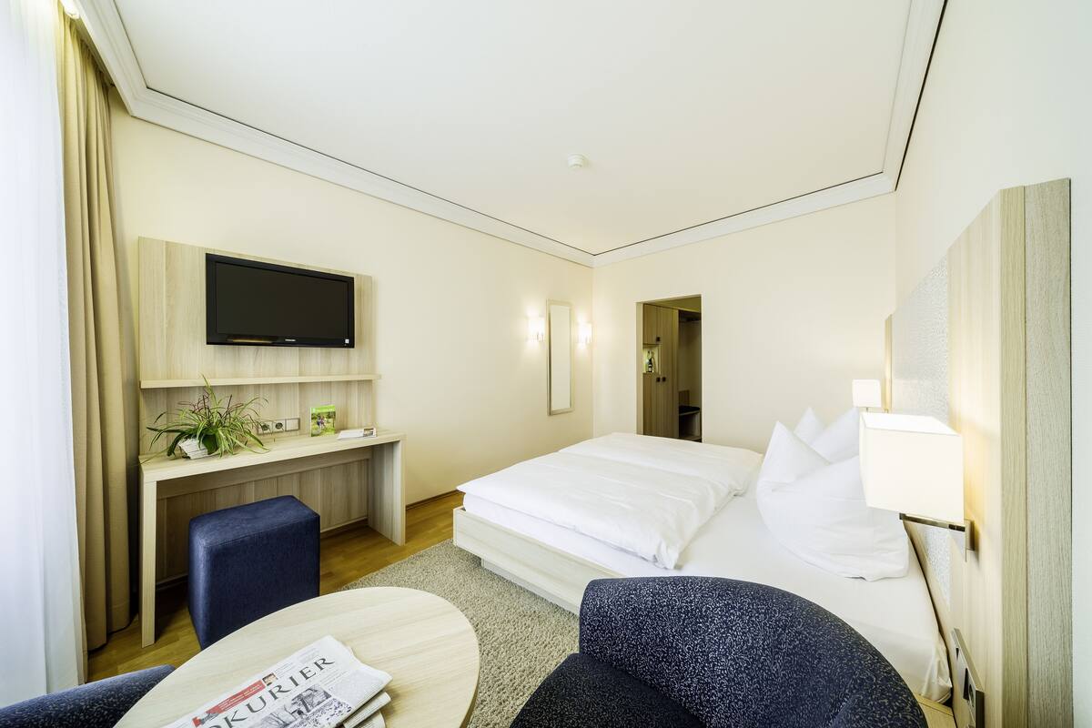 Een hotelkamer met een bed, een bureau met een televisie, een stoel, een klein tafeltje met een krant en een lamp.