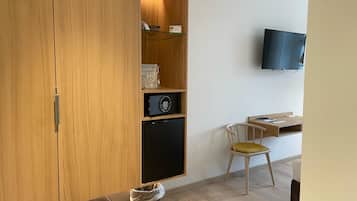 A modern room with a wooden wardrobe, a small black cabinet, a glass shelf with items, a mounted TV, a wooden desk, a chair, and a trash bin.