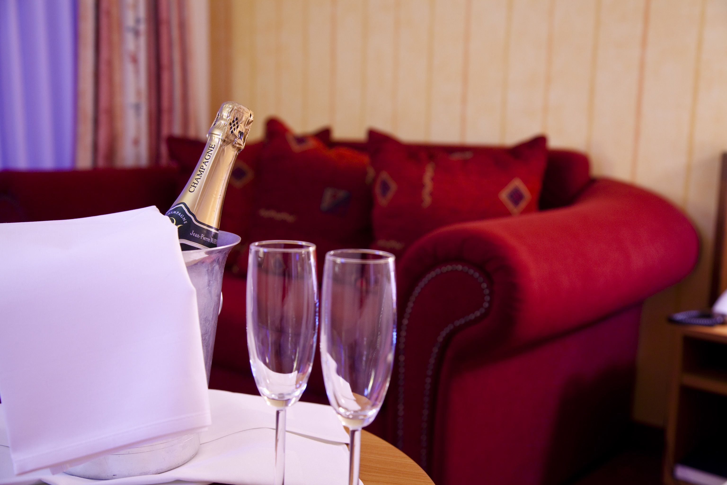 A table with a bottle of champagne and two glasses, a white napkin, and a red upholstered sofa in the background.