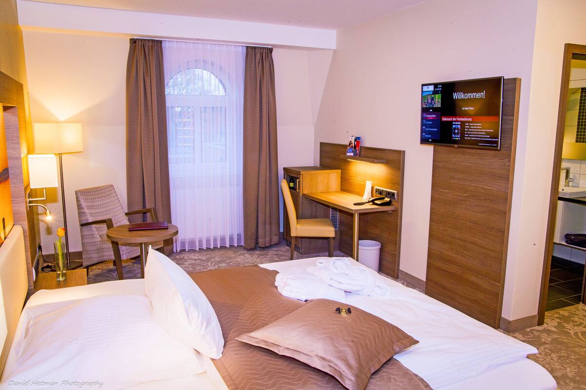 A hotel room with two beds, a desk, a chair, a lamp, and a television displaying a welcome message.