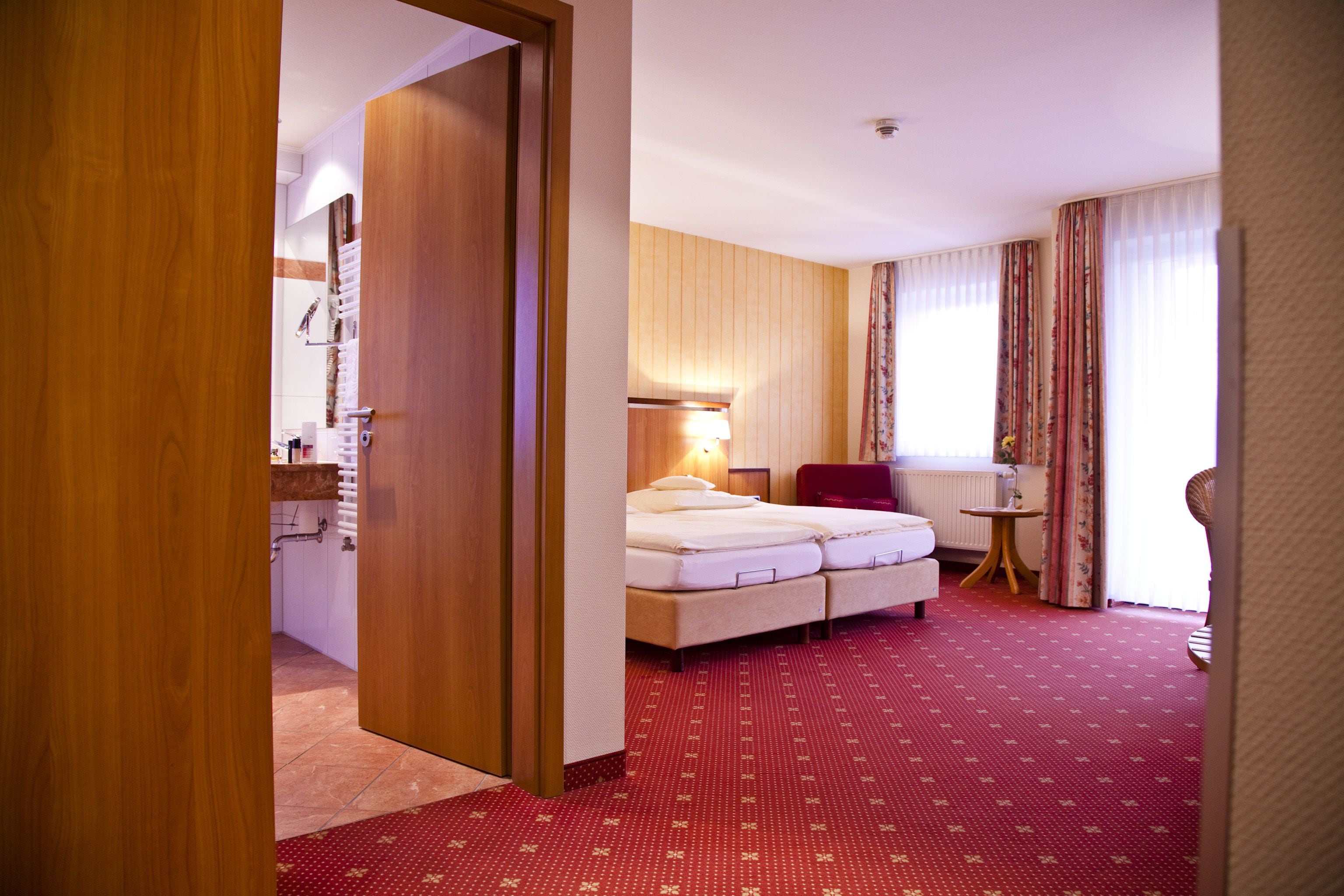 A hotel room with a wooden door, a bed with two pillows, a small table, and a chair.