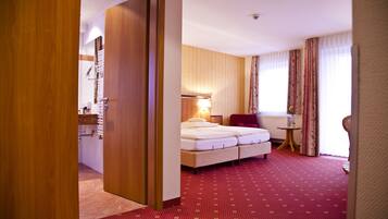A hotel room with a wooden door, a bed with two pillows, a small table, and a chair.