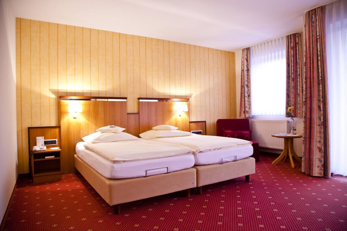 A hotel room with a double bed, two bedside tables, a chair, a small table, and a window with curtains.