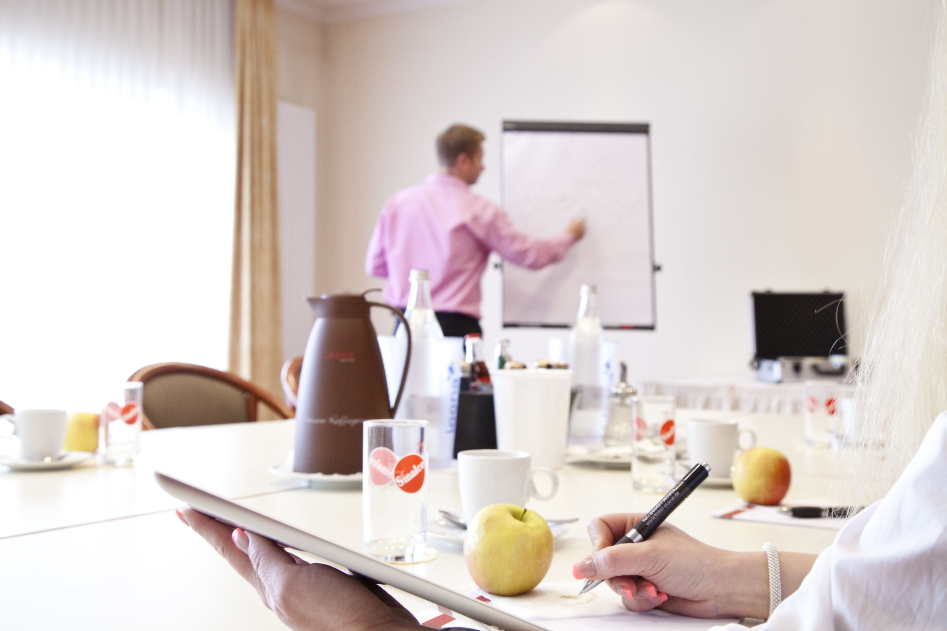 A conference room with a whiteboard, a coffee pot, a glass of water, and a tablet on the table.