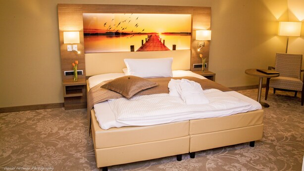 Habitación de hotel con una cama grande, dos lámparas de noche, una silla y un cuadro en la pared que representa un atardecer sobre un muelle.