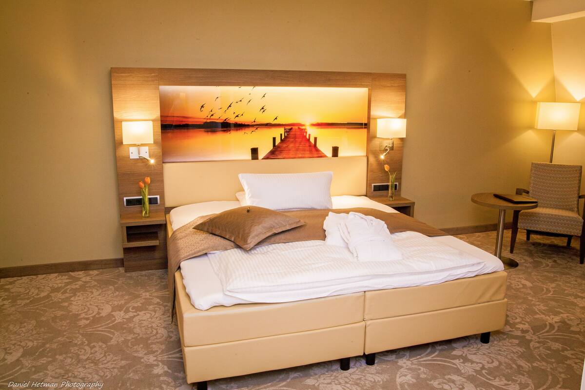 A hotel room with a large bed, two bedside lamps, a chair, and a wall-mounted painting of a sunset over a pier.