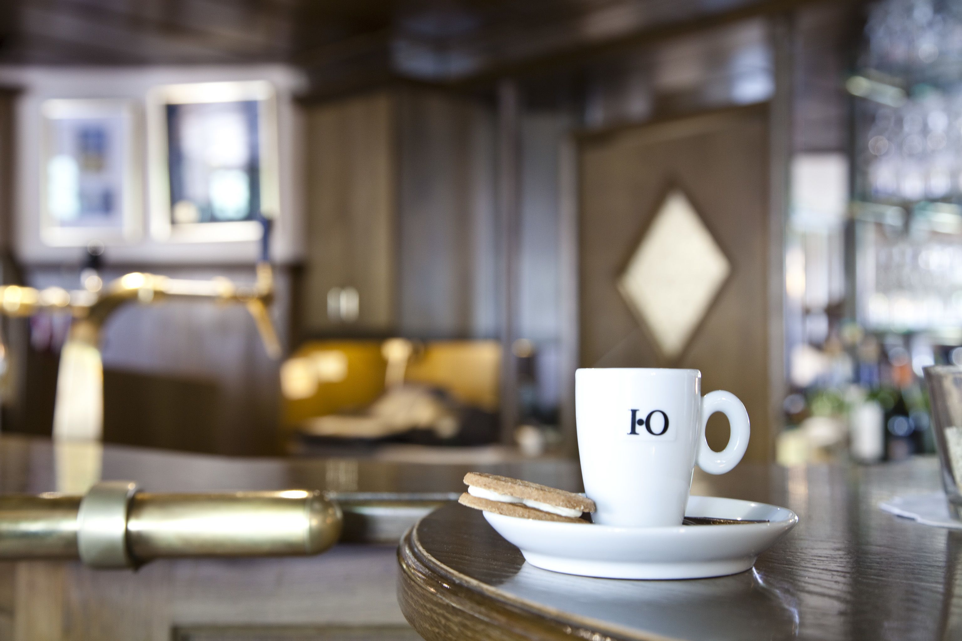 A white cup with a logo on a saucer, a sandwich, and a gold-colored object on a wooden table in a cafe setting.