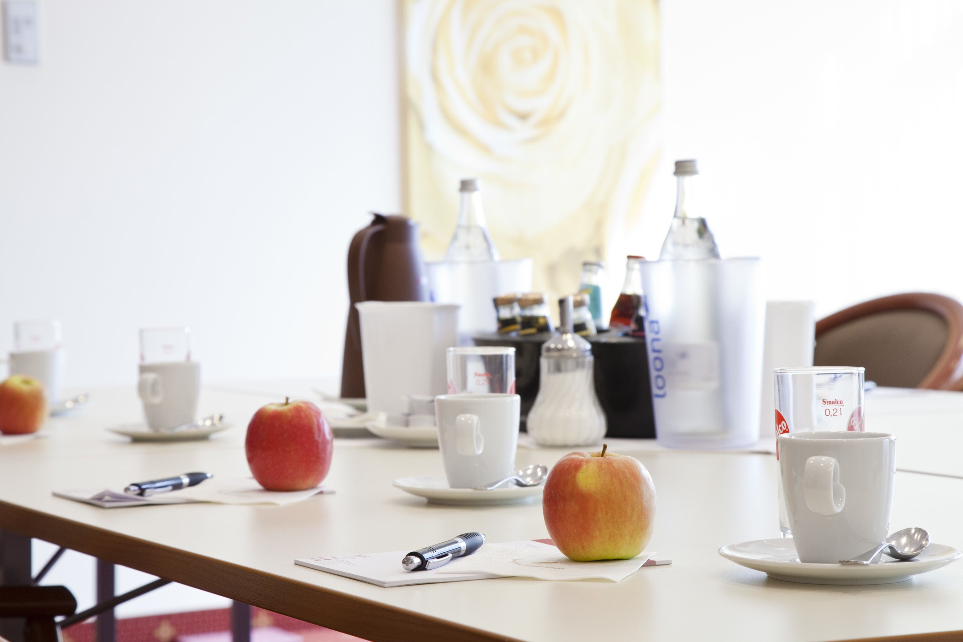 Ein Konferenztisch mit einer weißen Tasse, einem Glas Wasser, einem Löffel, einem Stift und einem Apfel.