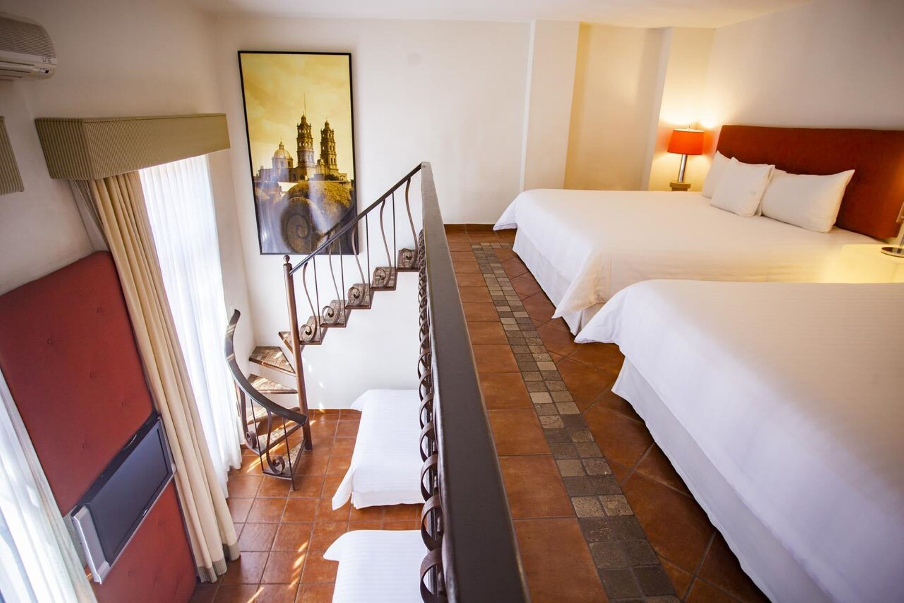 Habitación de hotel con dos camas, una escalera, un cuadro de un paisaje urbano y una pared roja.