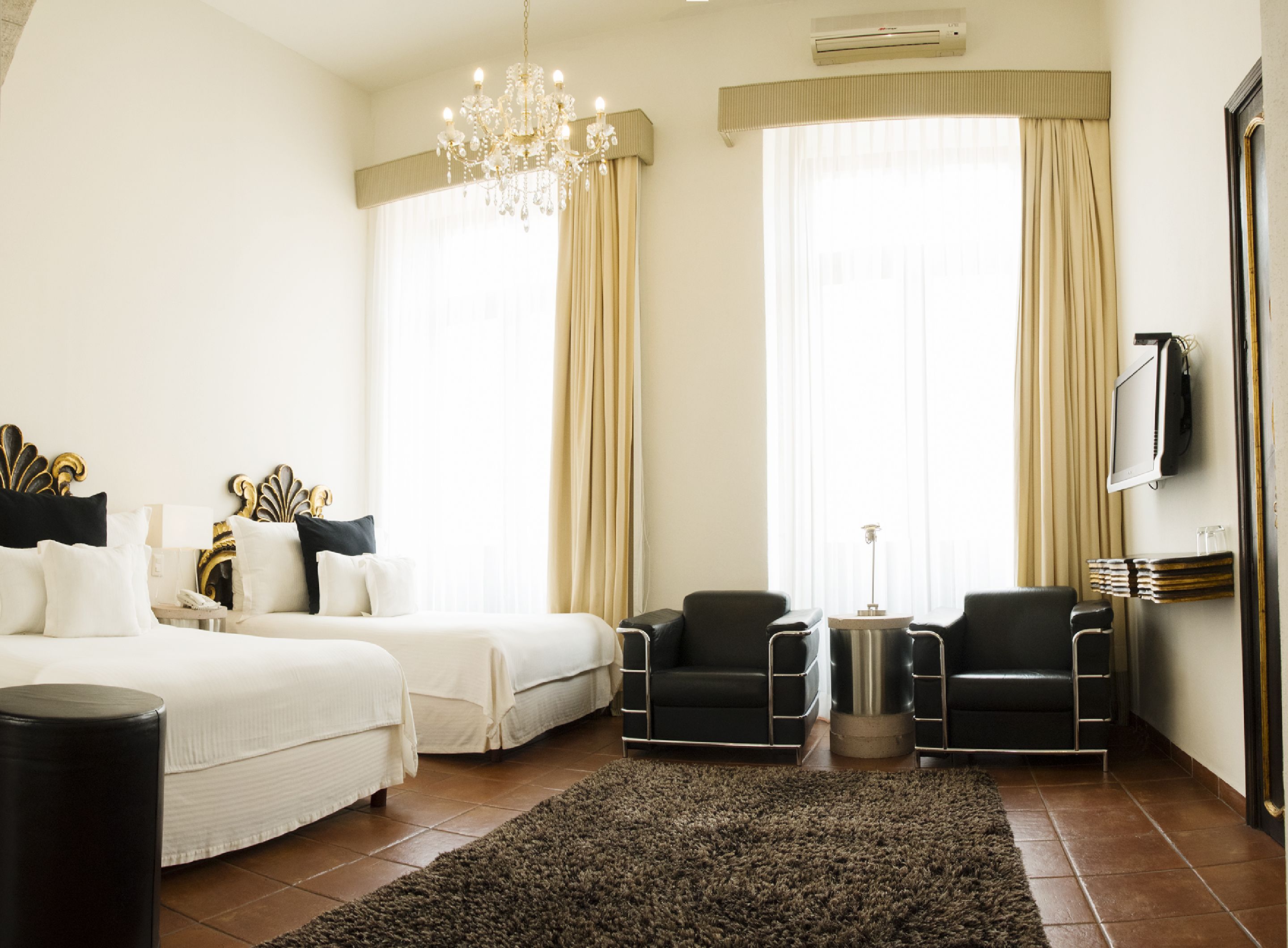 Une chambre d’hôtel avec un lit, deux fauteuils, une petite table, un lustre et un téléviseur fixé au mur.