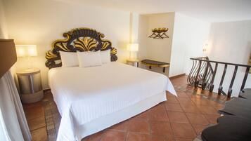Une chambre d’hôtel avec un grand lit, deux tables de chevet avec des lampes, une petite table, une chaise et un balcon avec une balustrade.