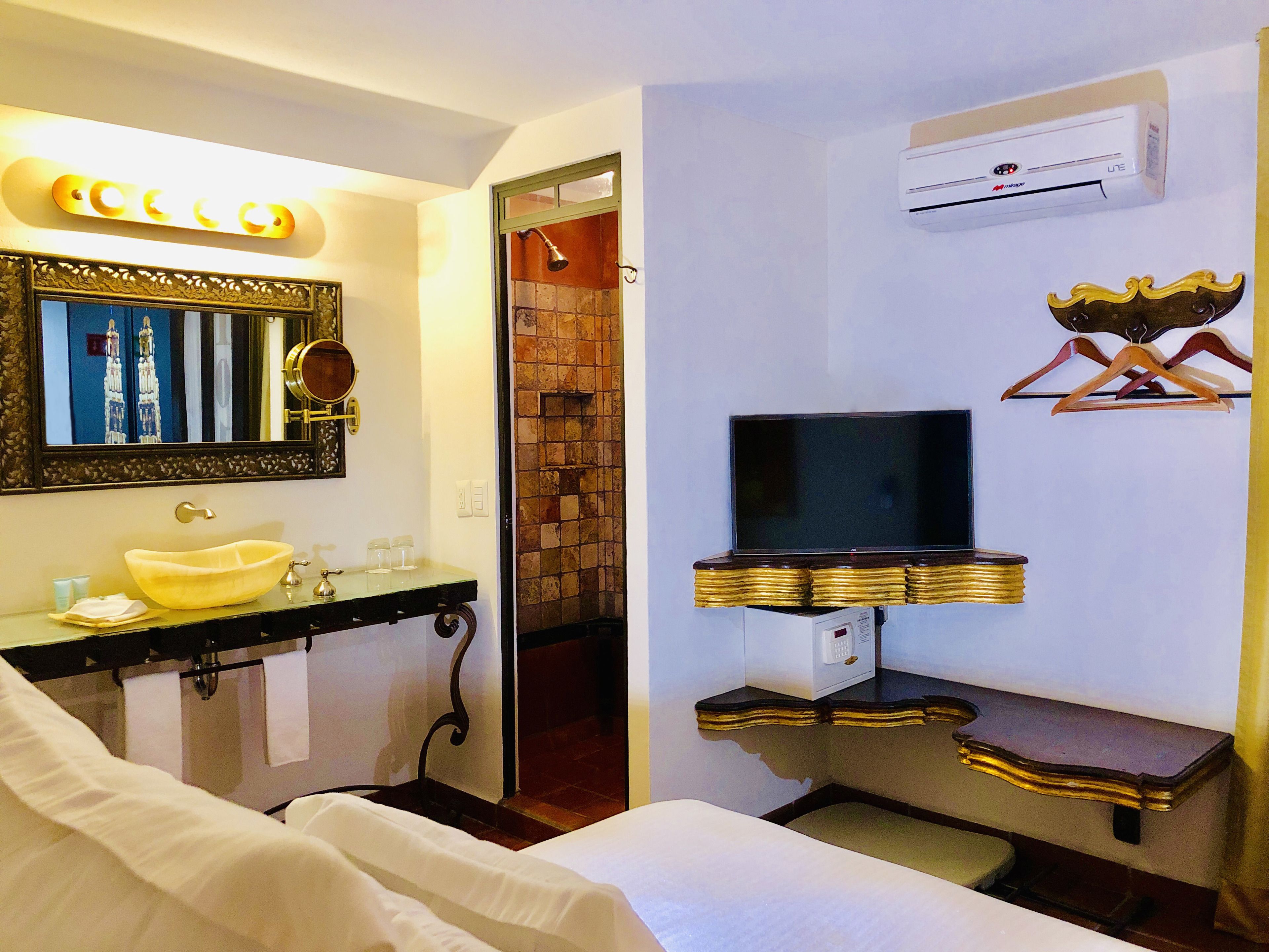 A hotel bathroom with a large mirror, a sink, a television mounted on a gold-framed shelf, and an air conditioning unit on the wall.