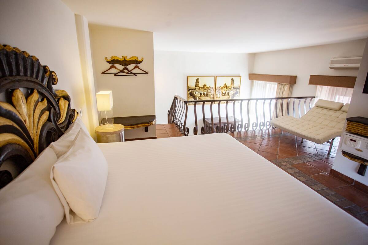 A hotel room with a large bed, a chair, a desk, a lamp, and a balcony with a view of a cityscape.