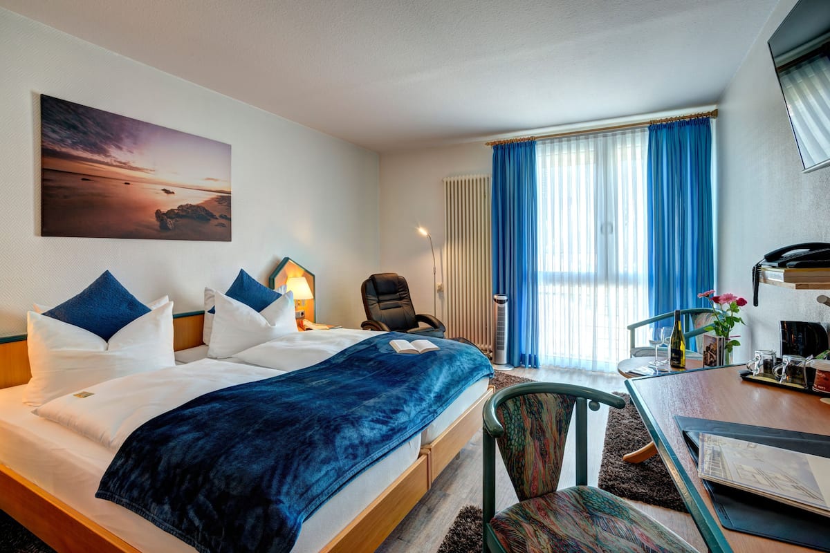 A hotel room with a bed, a desk, a chair, a lamp, a window with curtains, and a painting on the wall.