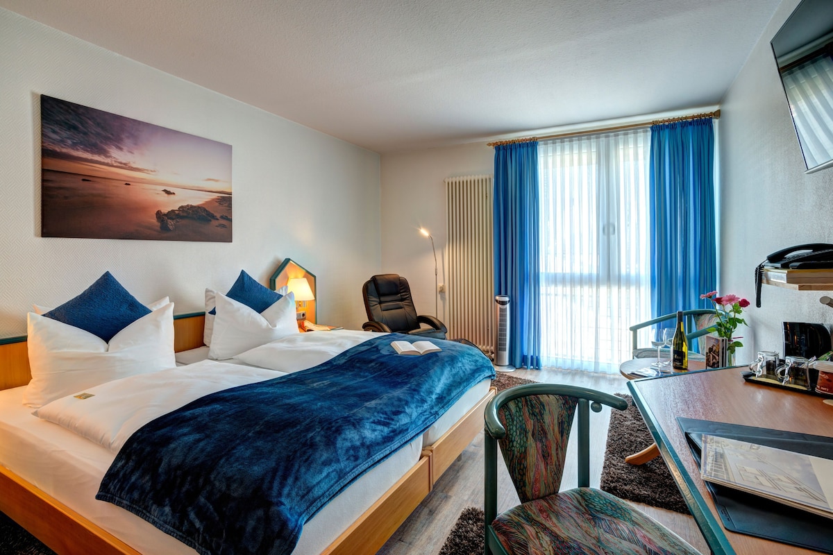 Habitación de hotel con cama, escritorio, silla, lámpara, ventana con cortinas y un cuadro en la pared.