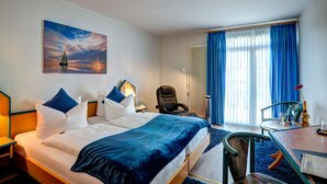 A hotel room with a bed, a desk, a chair, a lamp, a painting, and a window with curtains. - Hotel Paulin (Trier)
