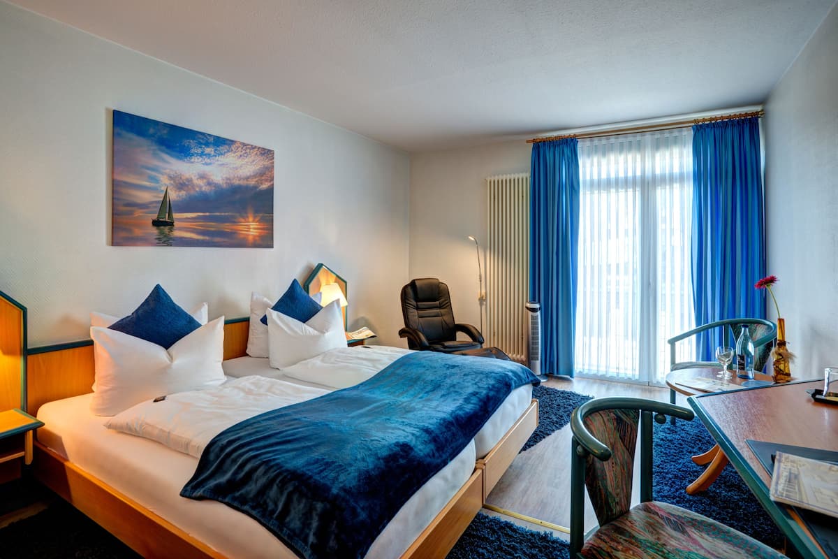 A hotel room with a bed, a desk, a chair, a lamp, a painting, and a window with curtains.