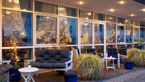 Un espace extérieur aménagé avec un canapé, des fauteuils et des tables, entouré de grandes fenêtres et de plantes en pot.