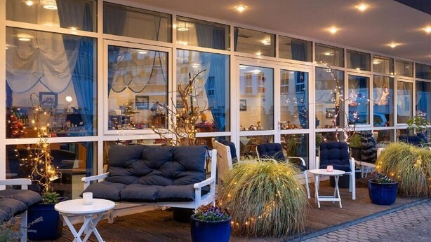 An outdoor seating area with a sofa, chairs, and tables, surrounded by large windows and potted plants. - Hotel Paulin (Trier)