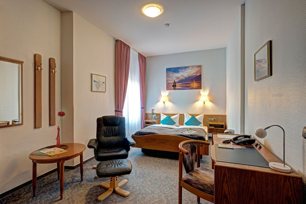 A hotel room with a bed, desk, chair, and a painting on the wall.