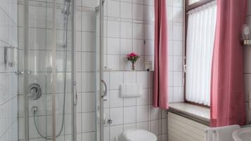 A bathroom with a glass-enclosed shower, a toilet, a sink, and a window with pink curtains.
