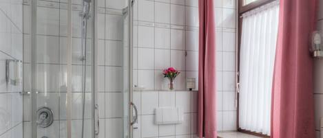 A bathroom with a glass-enclosed shower, a toilet, a sink, and a window with pink curtains.