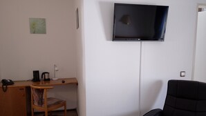A small, modern room with a flat-screen TV mounted on the wall, a wooden desk with a chair, a black armchair, and a small table with a kettle and phone. - Hotel Paulin (Trier)