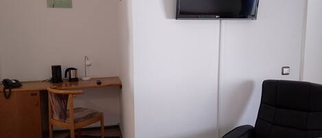 Una habitación pequeña y moderna con una televisión de pantalla plana en la pared, un escritorio de madera con una silla, un sillón negro y una mesita con un hervidor y un teléfono.