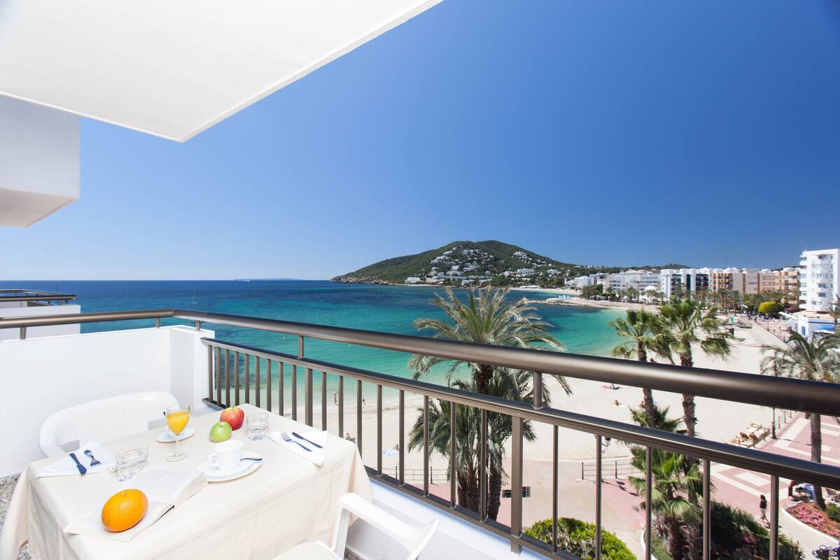 A balcony with a table set for breakfast overlooking a beach and the sea.