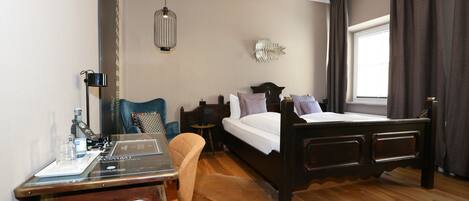Habitación de hotel con cama de madera, un escritorio con silla, una mesita, una lámpara y un amplio ventanal con cortinas.