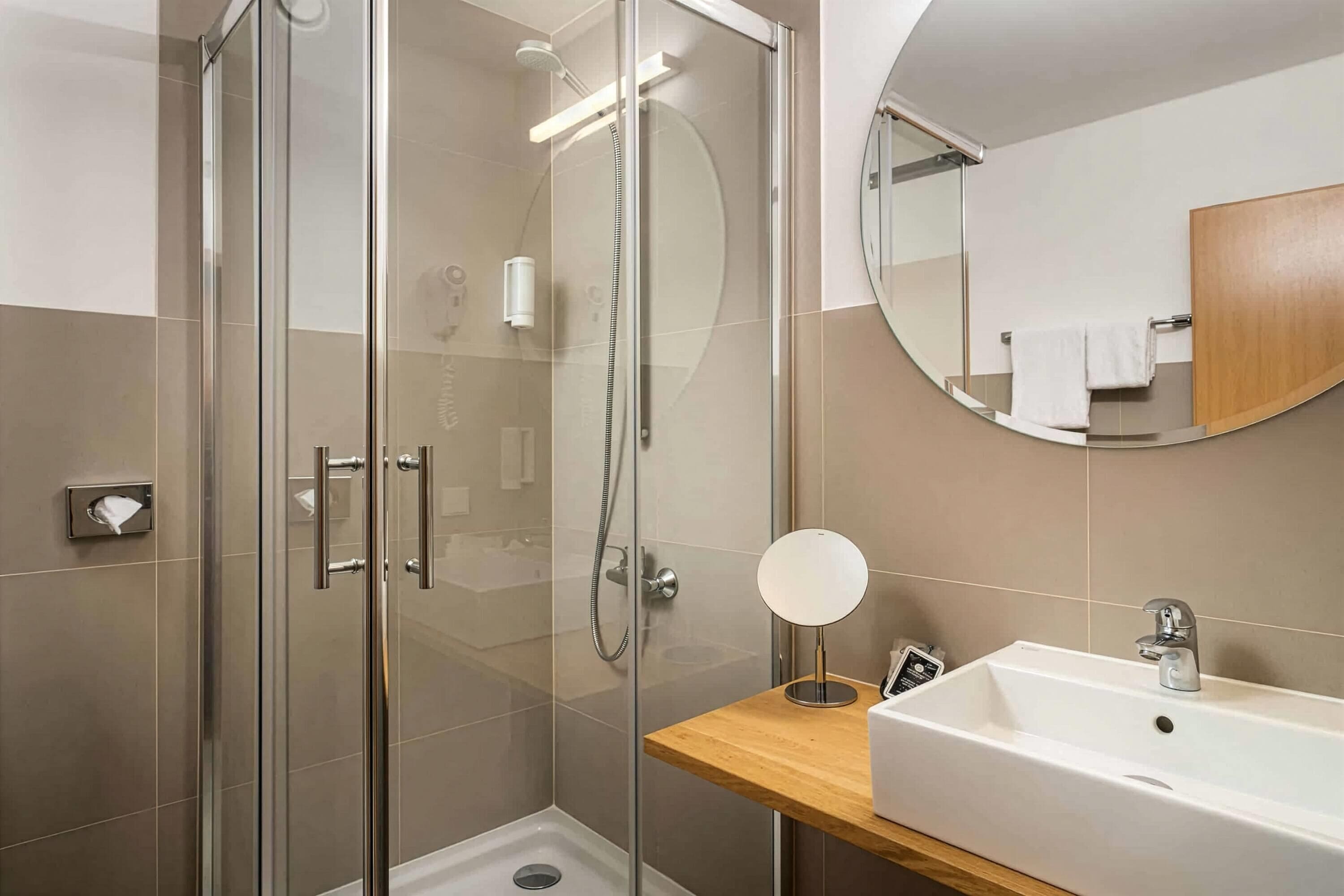 Un baño moderno con ducha de vidrio, tocador de madera con lavamanos y espejo redondo.