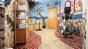 A room with a mosaic wall, a wooden shelf with towels, a door, and a stationary exercise bike. - Arthotel ANA Goggl, Trademark Collection by Wyndham (Landsberg am Lech)