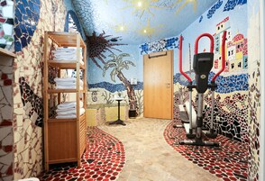 Quarto com parede em mosaico, prateleira de madeira com toalhas, uma porta e uma bicicleta ergométrica.