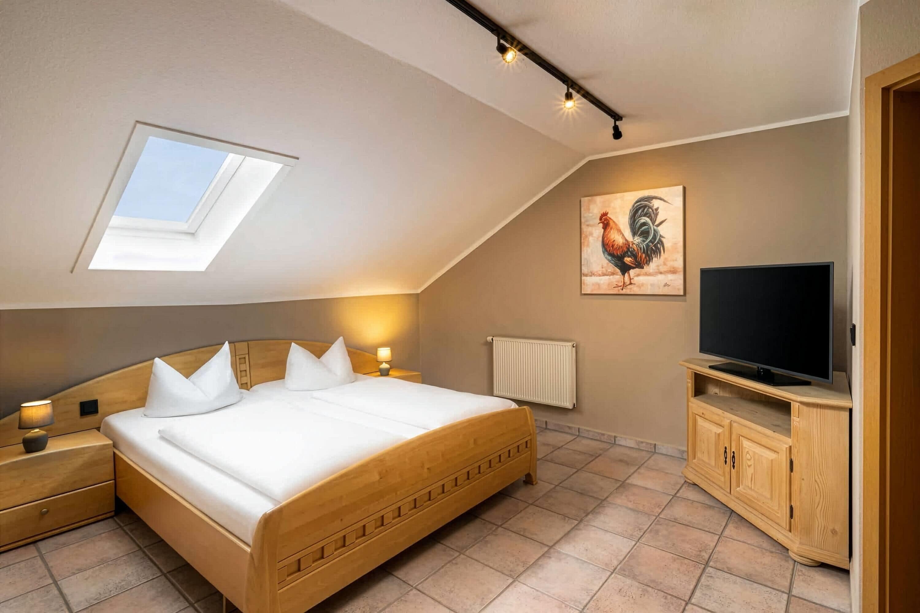 A bedroom with a sloped ceiling, a skylight, a bed with white bedding, a wooden headboard, bedside lamps, a radiator, a television on a wooden cabinet, and a rooster painting on the wall.